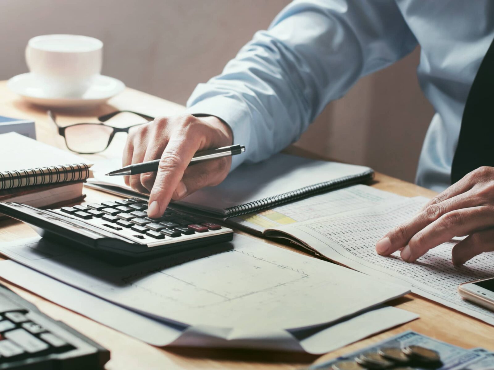 A person in a blue dress shirt and tie uses a calculator and pen to examine detailed figures on paper, surrounded by notebooks and money.