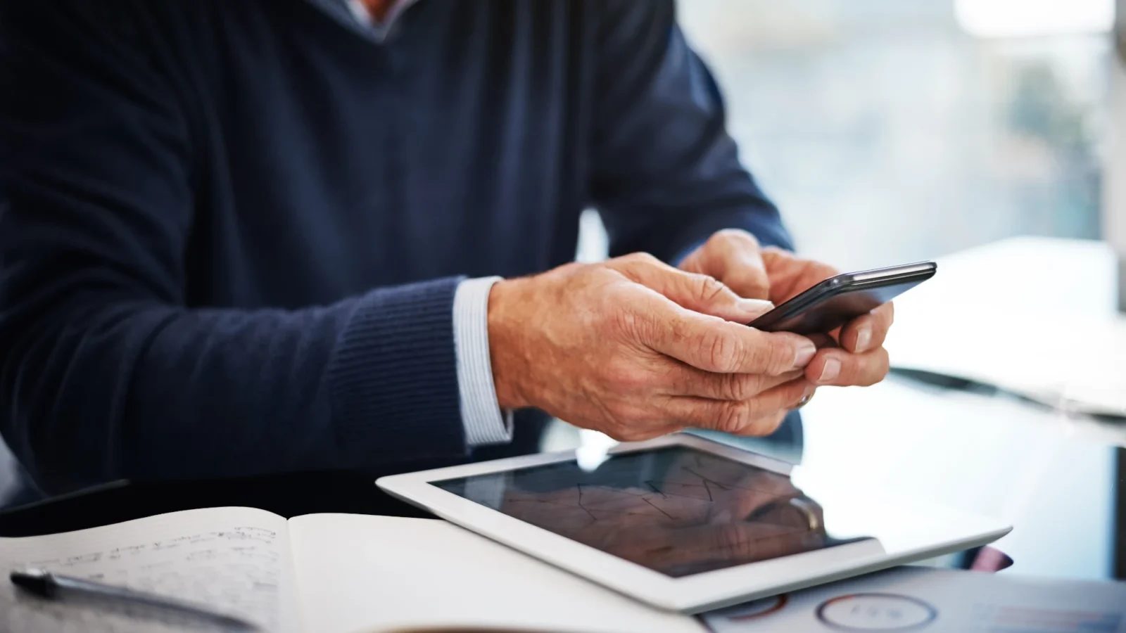 A person in a dark blue sweater uses a smartphone over a desk that holds a tablet and printed documents displaying business charts and graphs.